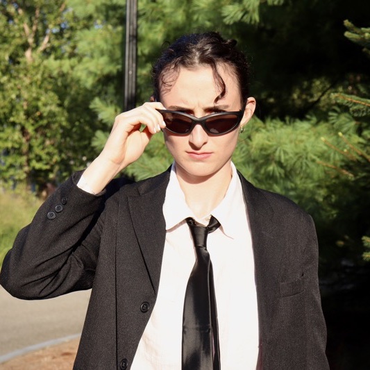 A portrait of Noe Rueschemeyer Bailey, a person with short brown hair, sunglasses, a black coat, and a white shirt.