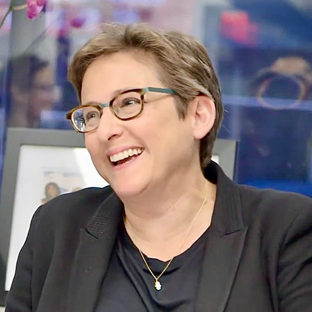 Rabbi Sharon Kleinbaum smiling, wearing a gold chain, black shirt, black jacket and eyeglasses.