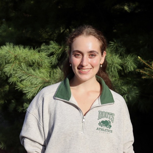 A portrait of Sarra Grimshaw, a person with long brown hair and a gray hoodie that says "Roots Athletics" on it.