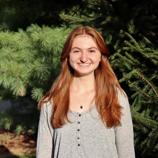 A portrait of Sophia Maron Schaeffer, a person with long red hair and a gray shirt.