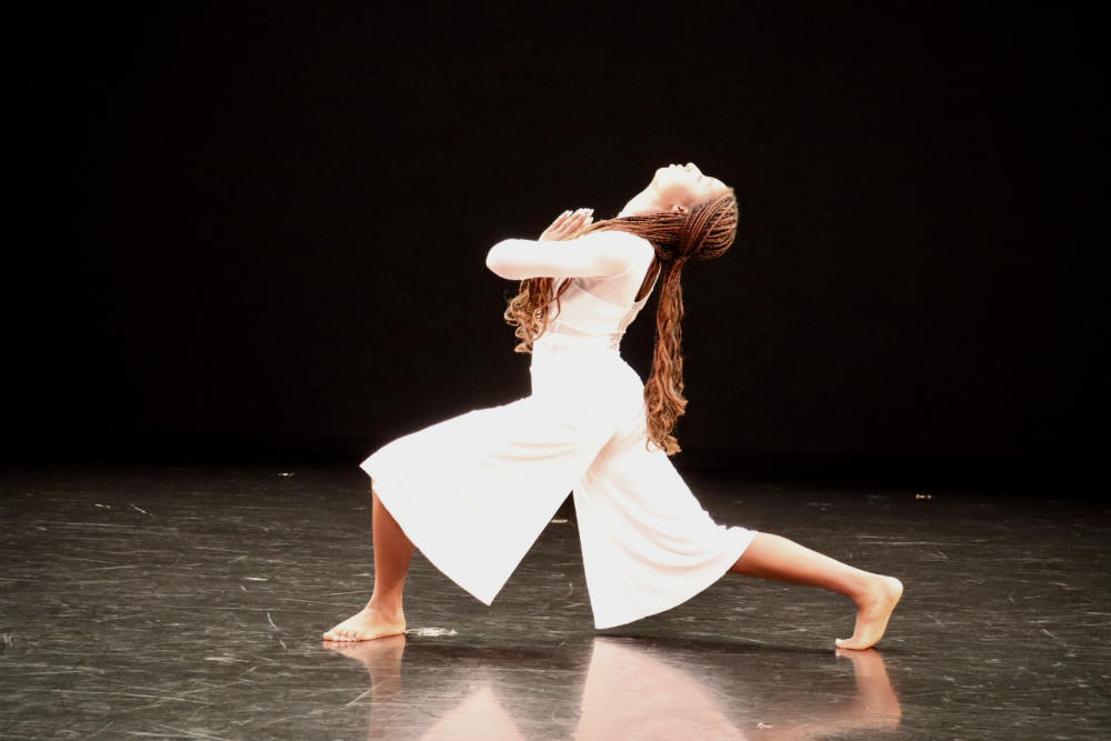 A person in a white dress and long dark hair performs on a dark stage.