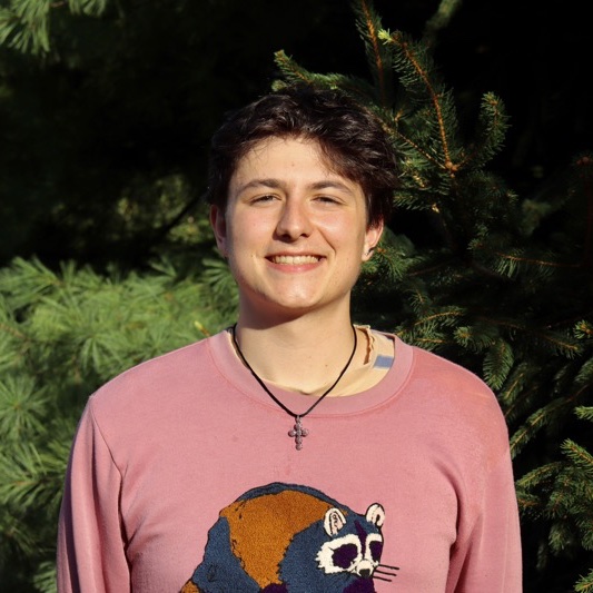 A portrait of Zack Garipoli, a person with short brown hair and a pink shirt with an illustration of a raccoon on it.