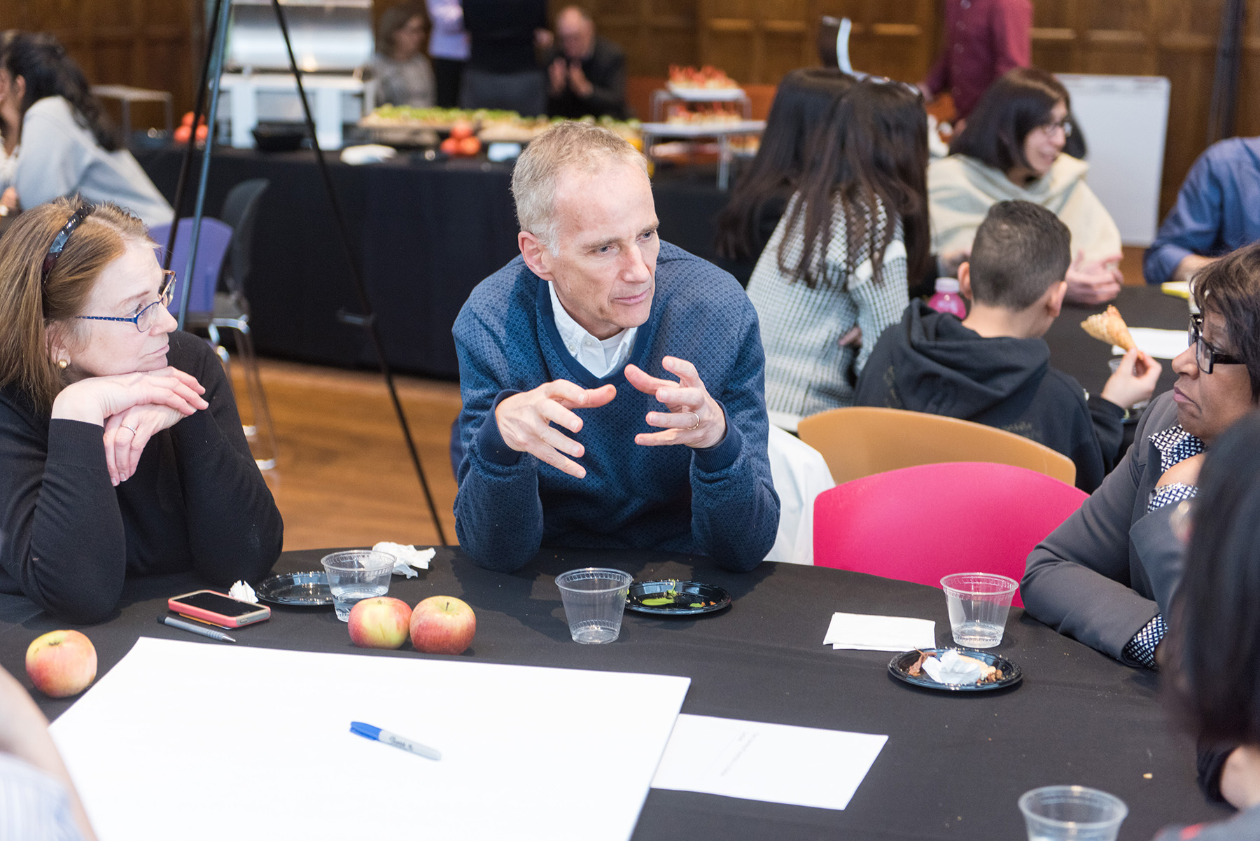 Person sitting at a round table speaking to the people sitting next to them. There are other tables with people sitting around them in the background.