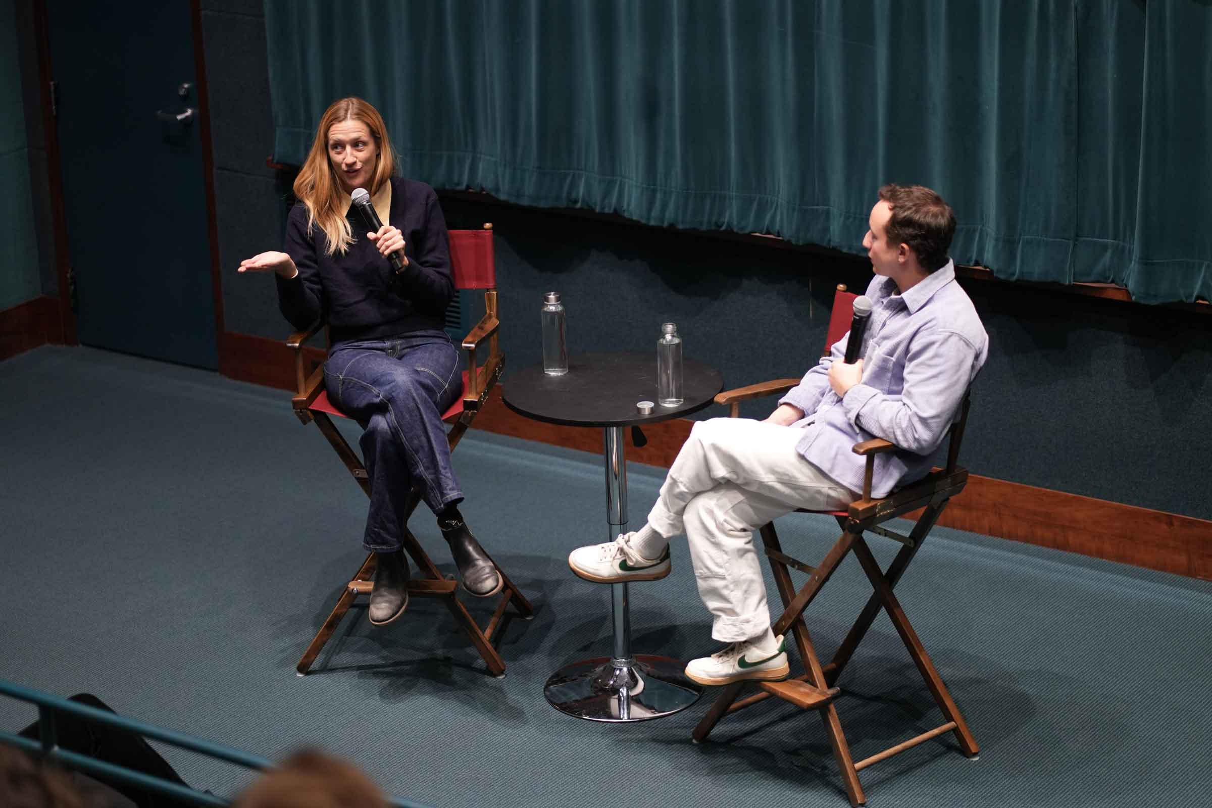 Two people seated in directors chairs on a stage holding microphones having a conversation.