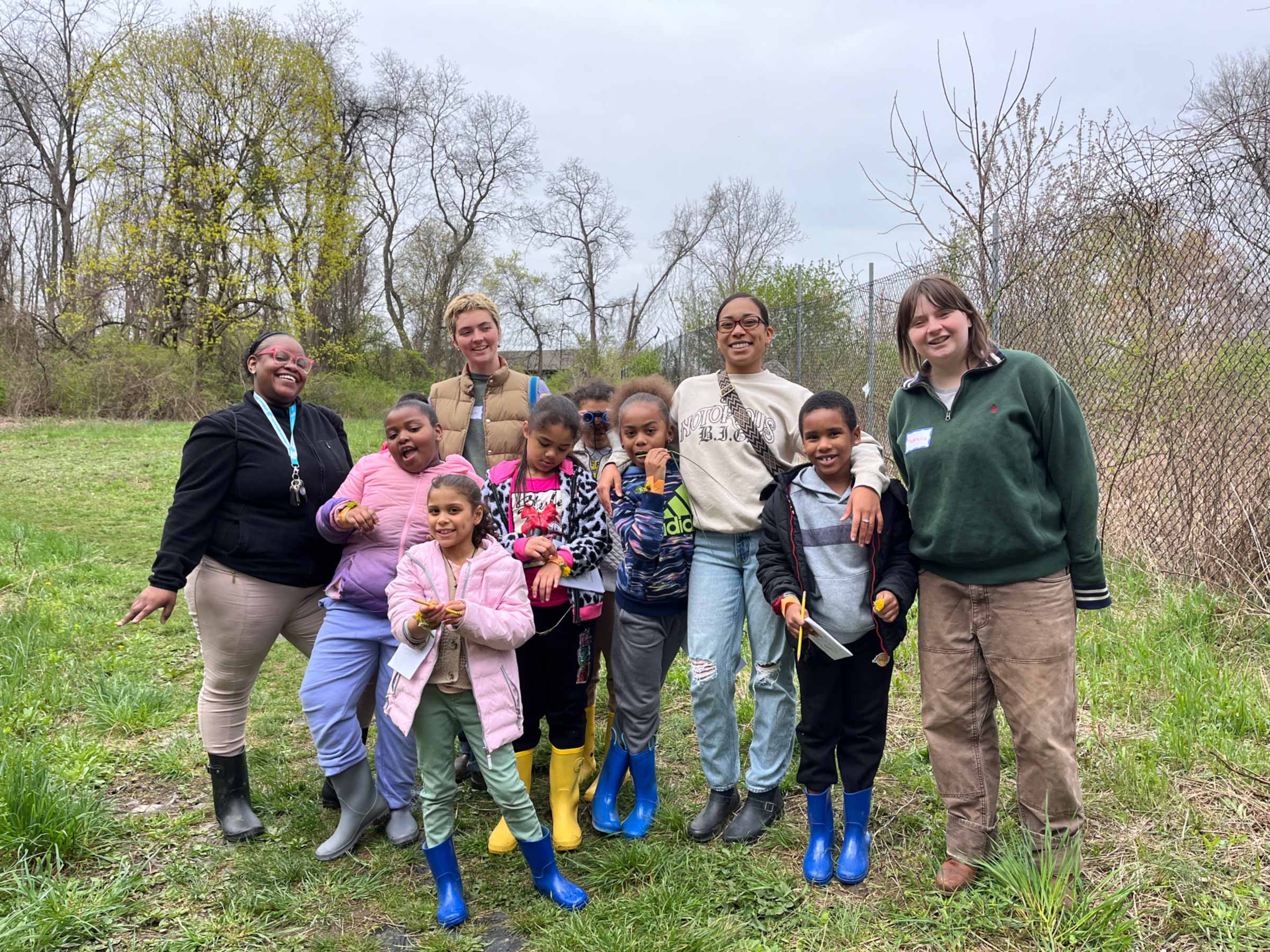 Group of adults and children in work boots and clothes standing in a field with trees in the background.