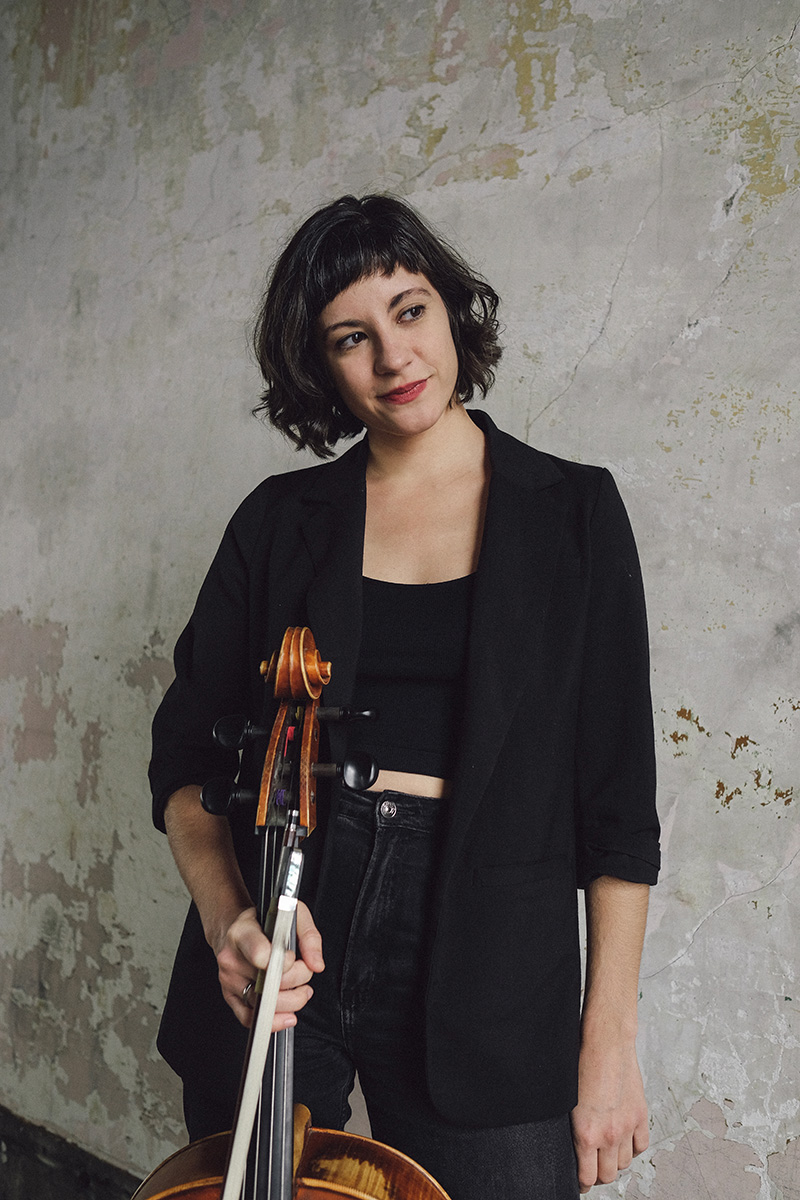 Person with shoulder-length dark hair standing next to a cello with their head tilted to the side looking away to the right.