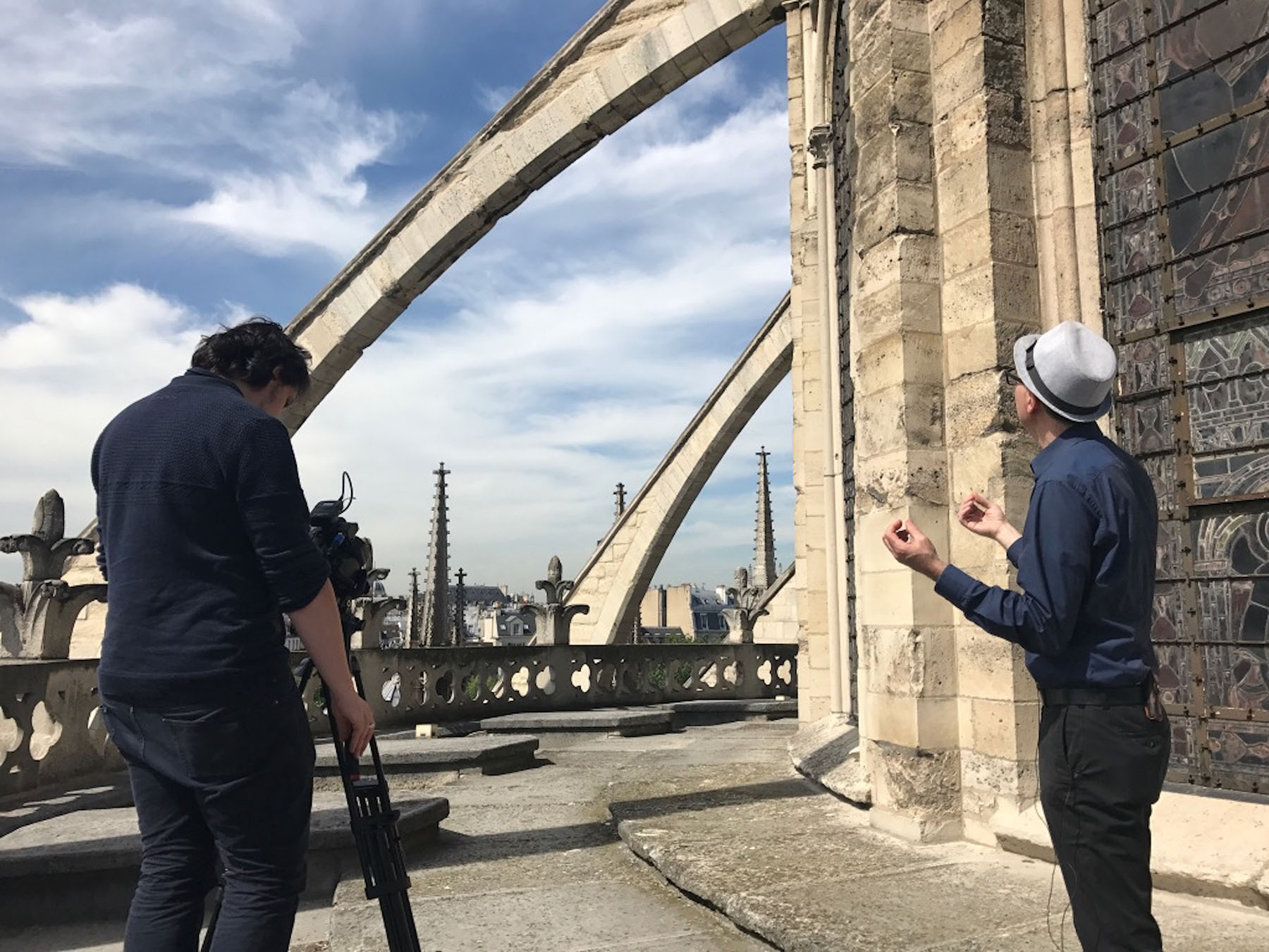 A person, from the back, wearing all black in front of a camera on a tripod and another person wearing a navy blue shirt, black pants and white hat with both arms out in front facing a stone building.