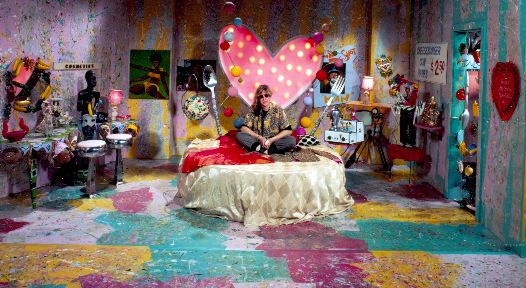 Allee Willis sits on a circular bed in the middle of a music video set, the walls are splatter painted and there is a heart with lights surrounding it on the wall behind her.