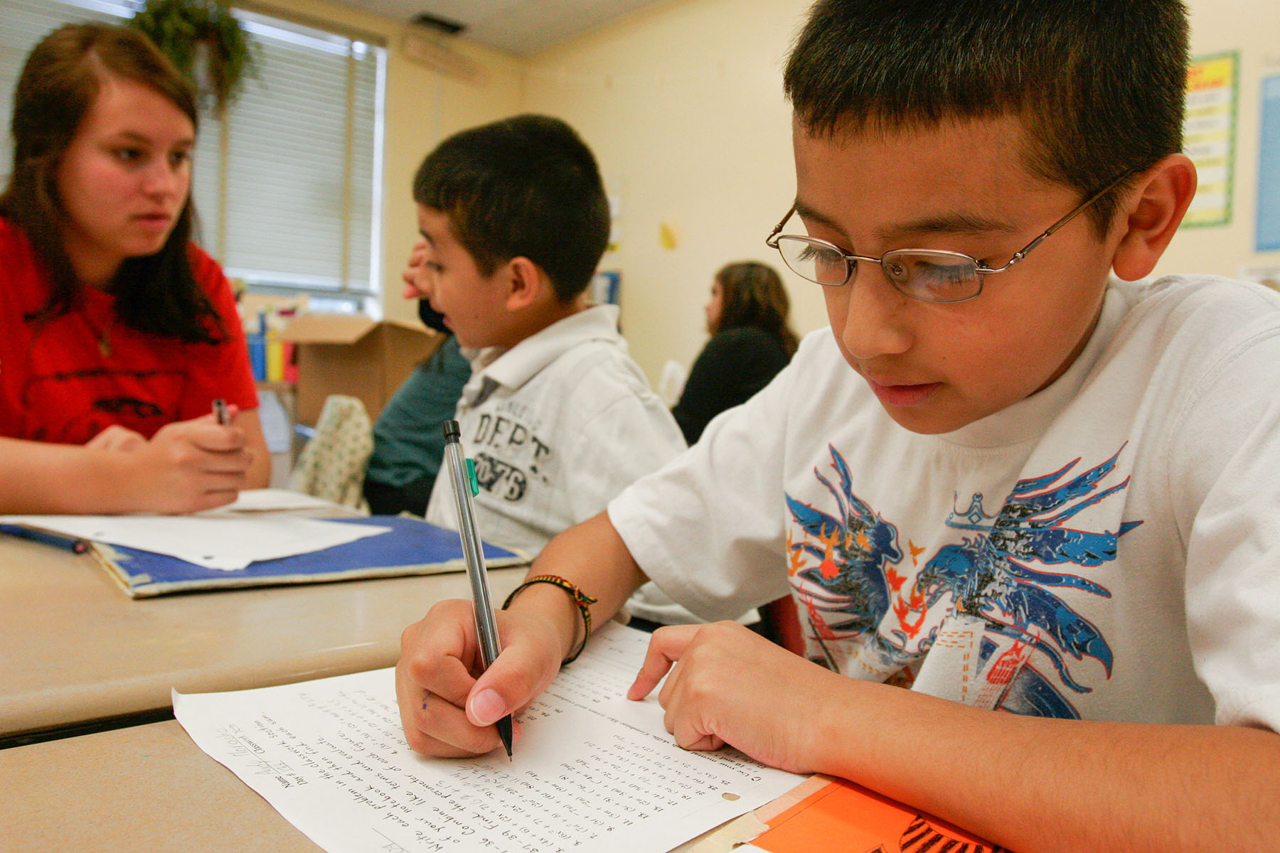 A person sitting at a table writing on a piece of white paper while a teacher and student speak in the background.