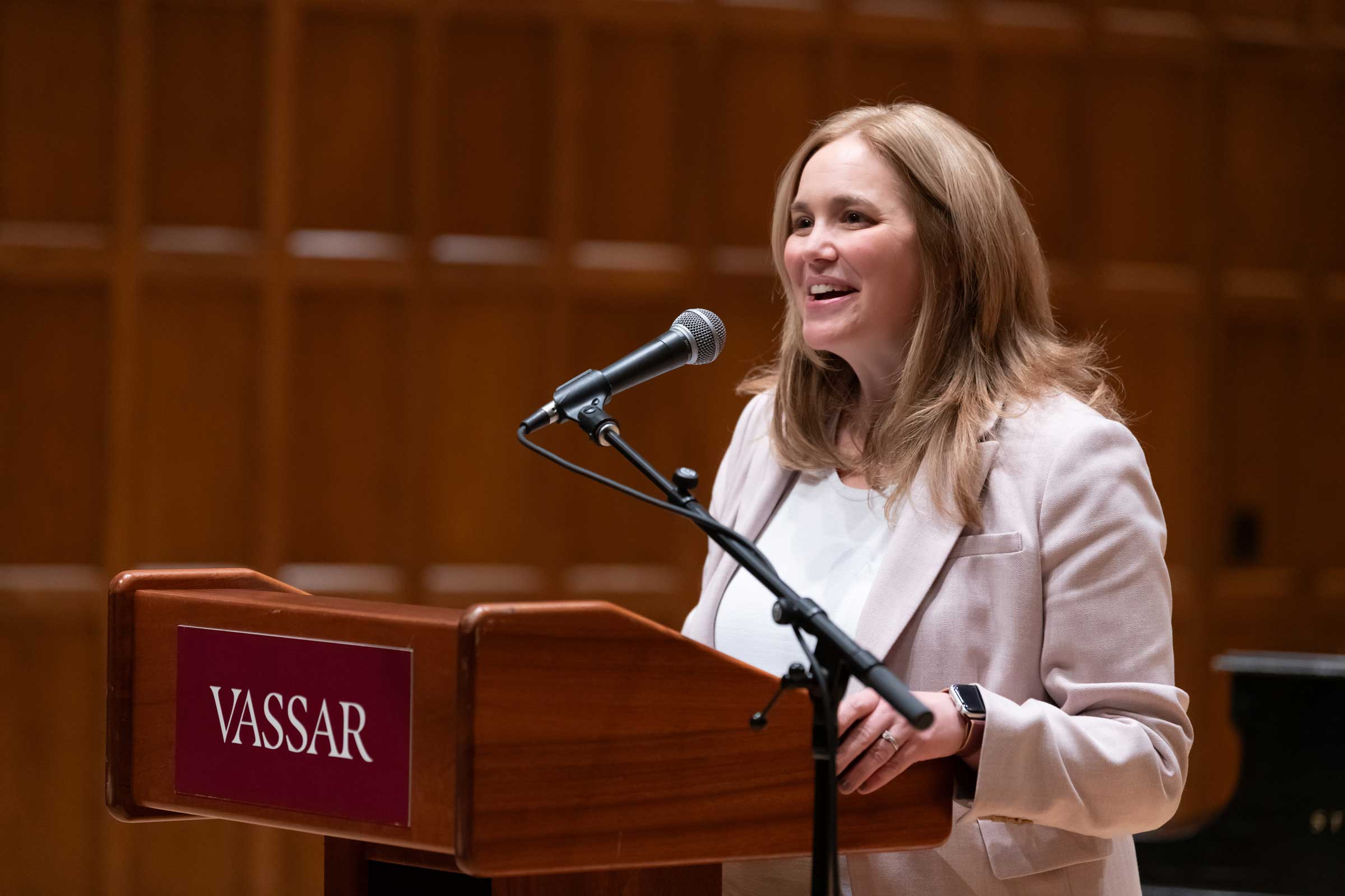 A smiling person behind a lectern with a microphone addressing an audience off camera.