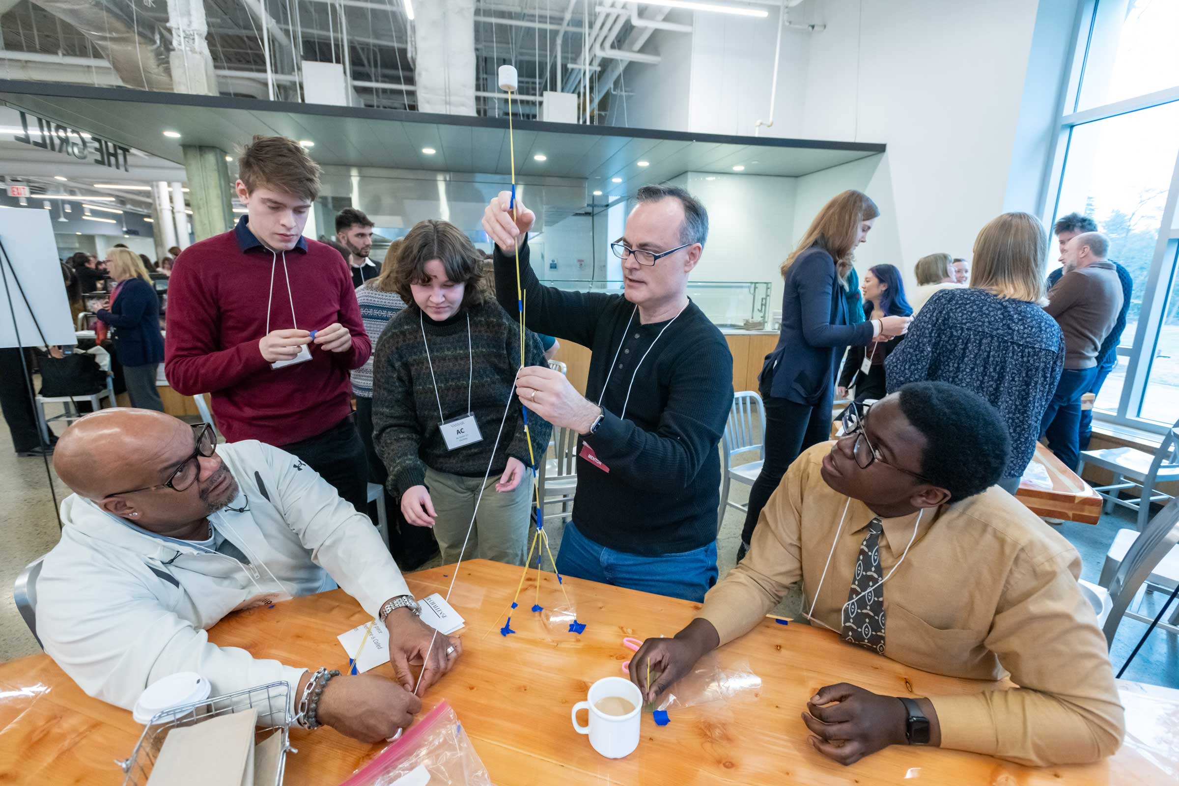 People seated around a table watching a standing person build a tower out of spaghetti noodles, tape, marshmallows.