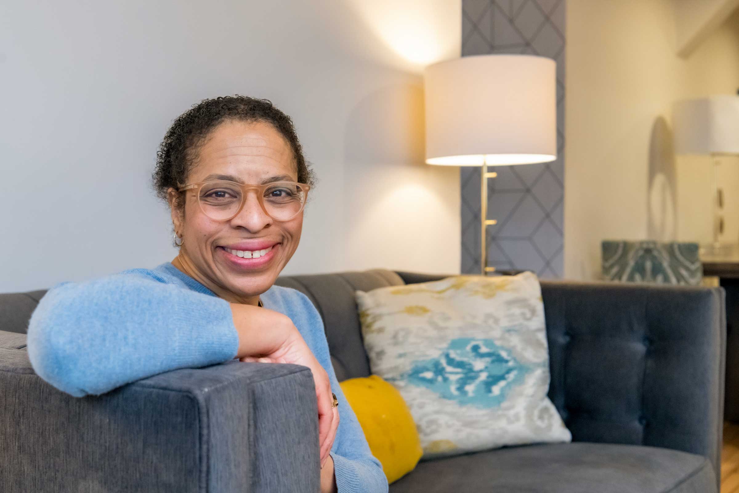 Photo of a smiling person sitting on a couch looking over the armrest.