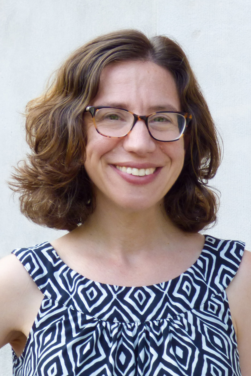 A person with glasses and shoulder-length red/orange hair wearing a blue and white diamond-pattern sleeveless top smiling for the camera.