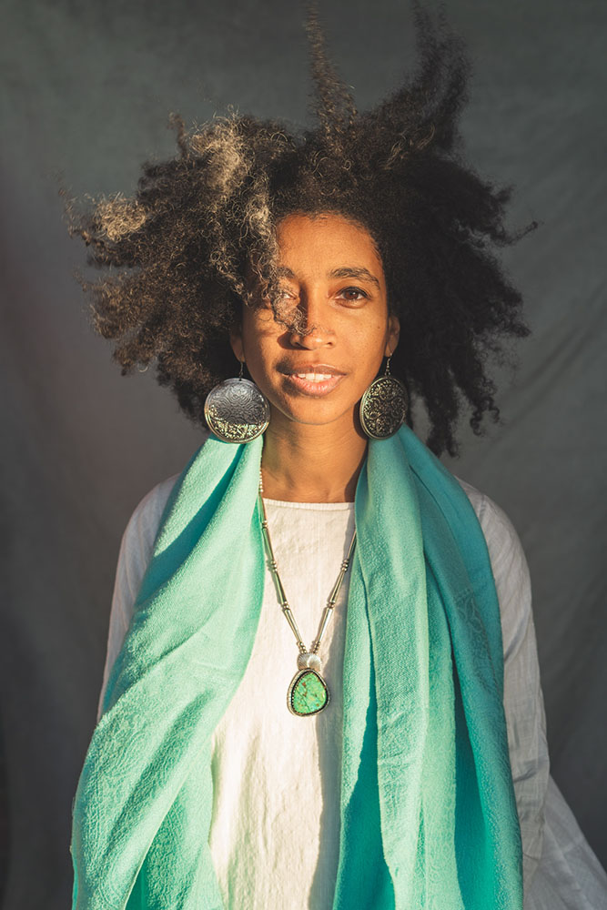 Person wearing a white long sleeve shirt, long teal scarf, necklace with large teal stone, large round gold earrings with long dark curly hair. 