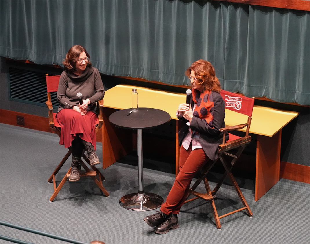 Professor Dara Greenwood and Prudence Fenton sitting on director style chairs holding microphones with a small, tall round table between them.