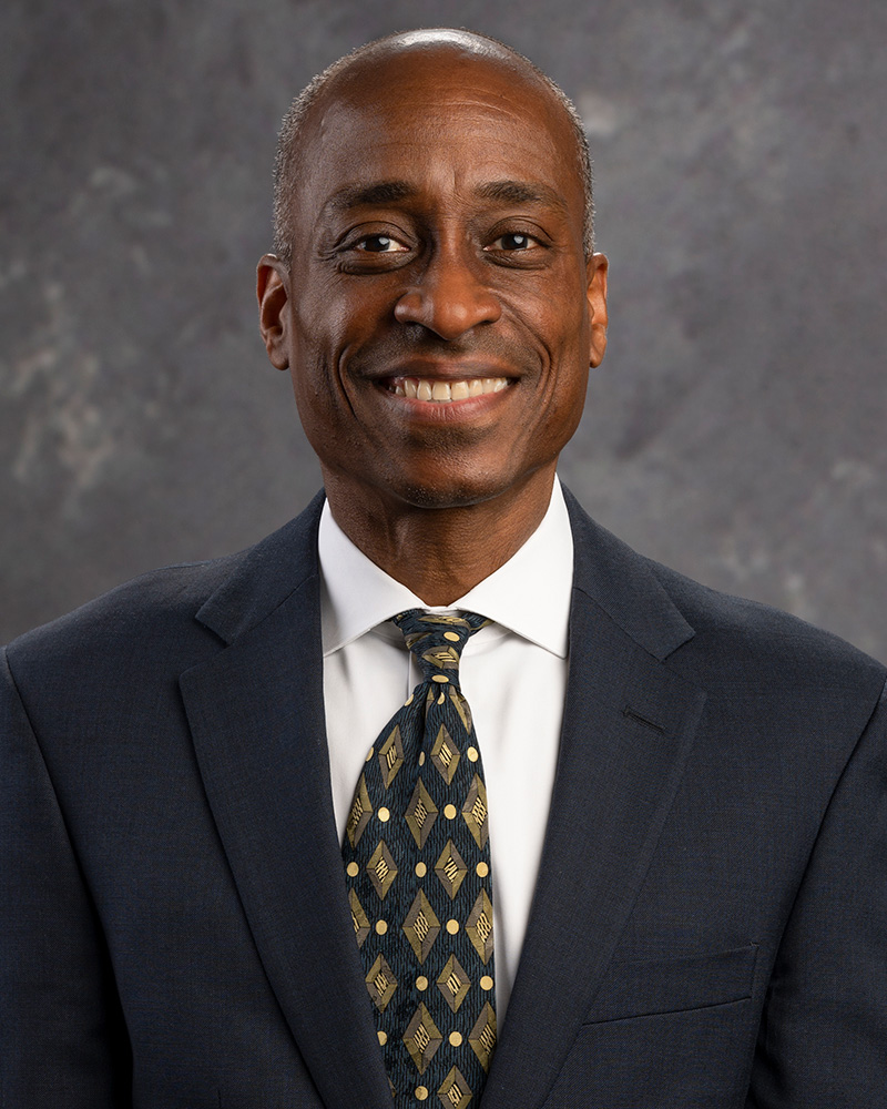 Portrait of a person smiling in a dark blue navy suit jacket, white shirt and blue and gold tie.