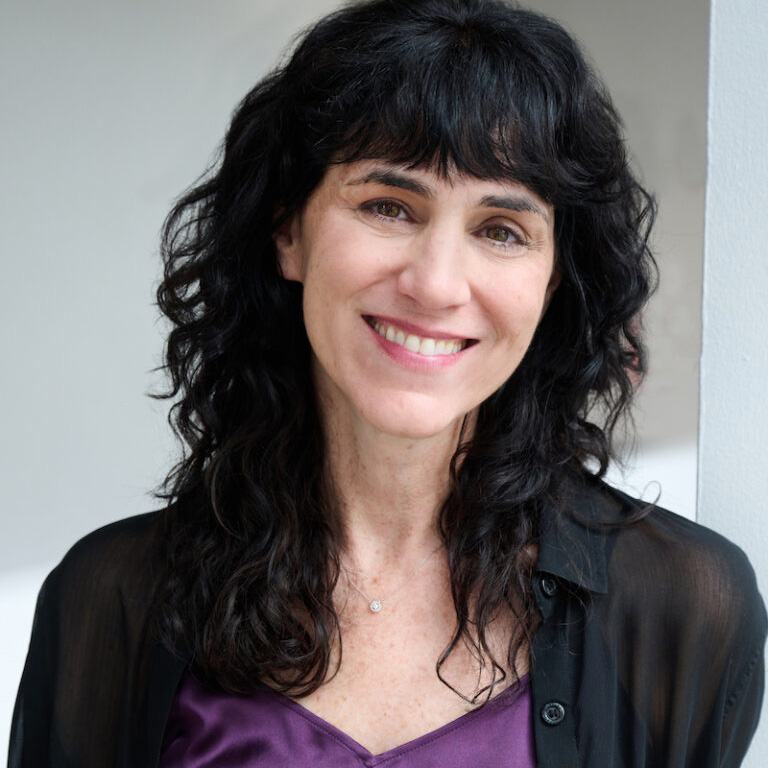 Leigh Silverman with long curly dark hair wearing a purple shirt under a black unbuttoned collared shirt. 