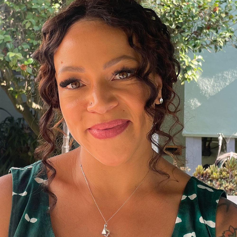 Person smiling wearing a green no sleeve top with white spots and curly dark hair pinned up and a tree in the background.