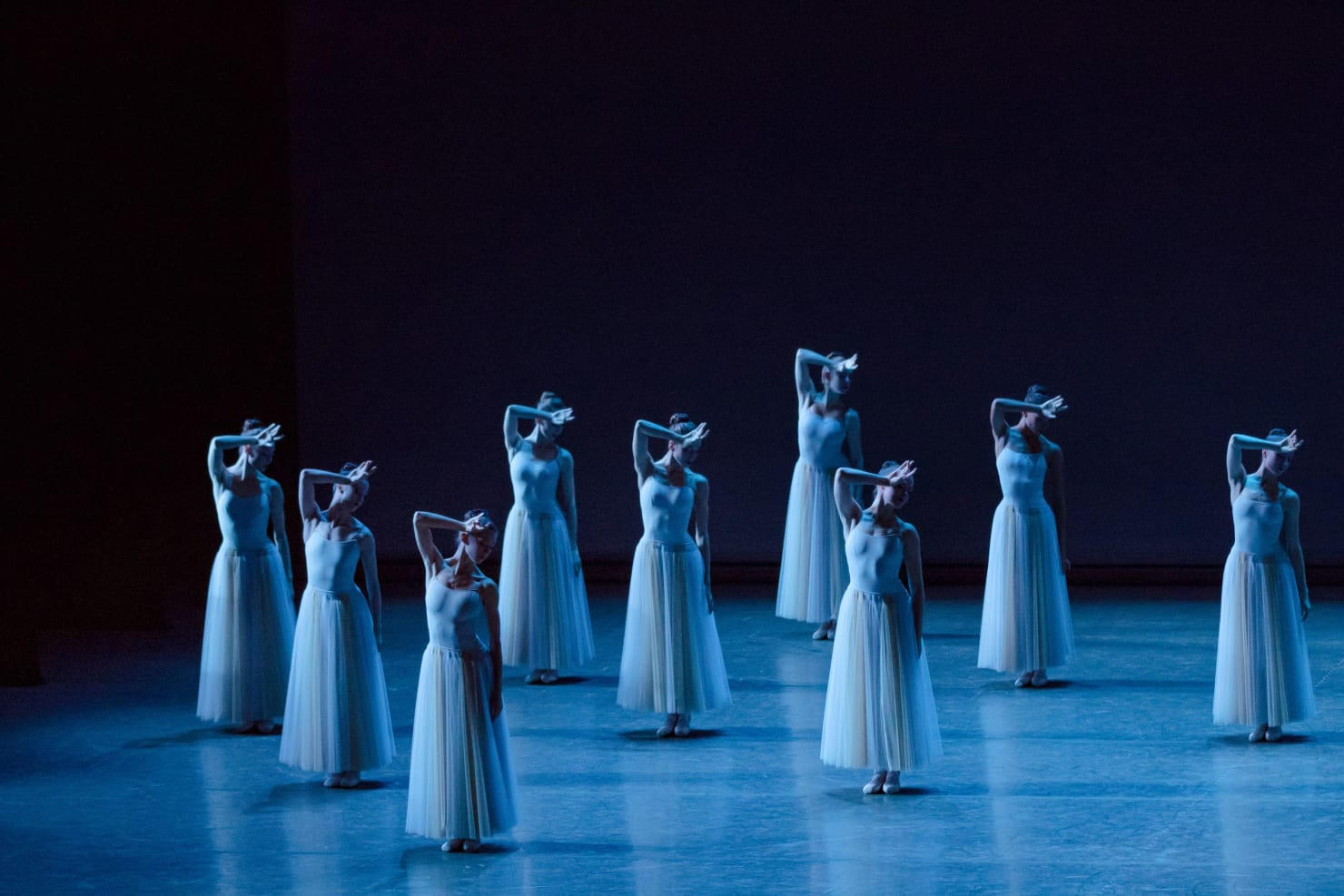 Many people in long white dresses perform on a darkened stage.