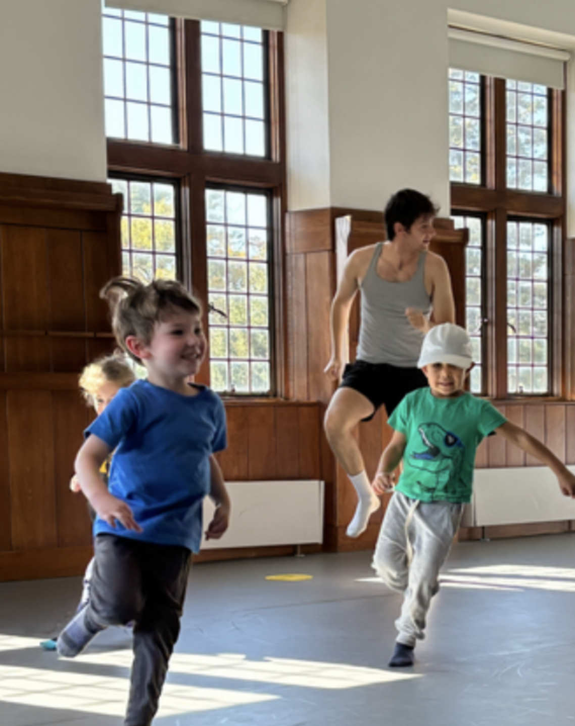 A bright room with large windows. Several children dance in the room. An adult stands behind them.