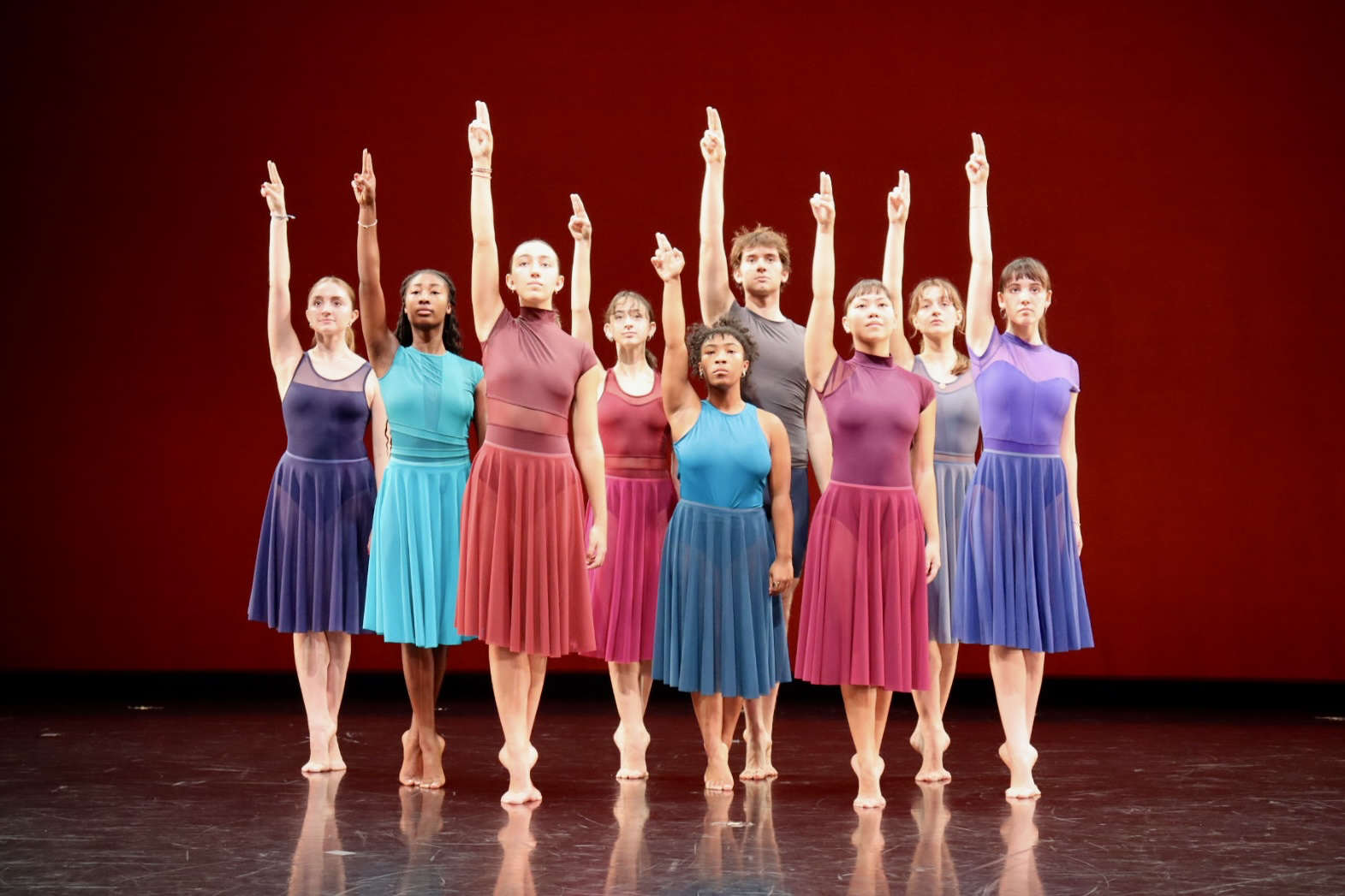 A group of people wearing brightly colored clothing stand on stage, right arms held straight up.