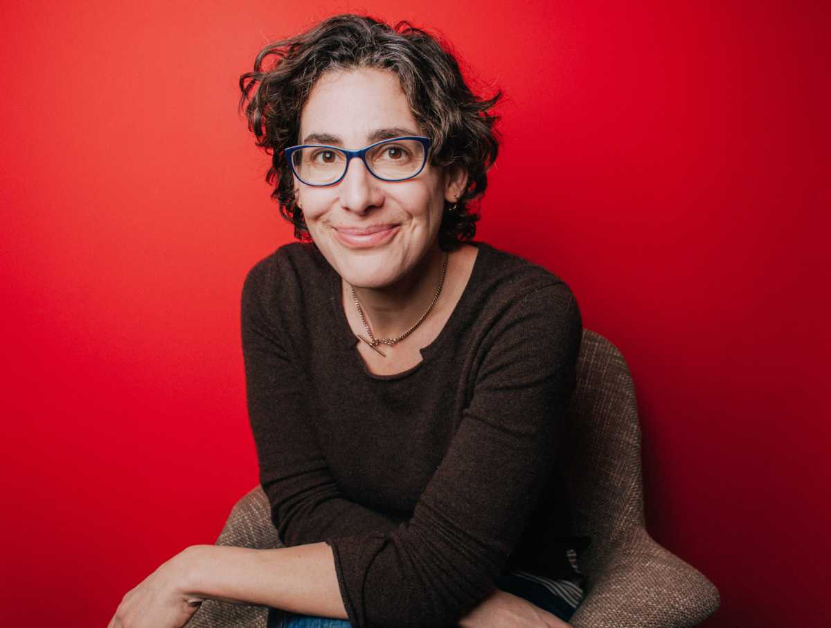 A portrait of Sarah Koenig, a person with medium-long brown hair, a dark sweater, and glasses. Koenig is sitting against a red background.