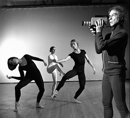 A grayscale photo showing three people performing dance moves while a fourth person films the scene with a video camera.