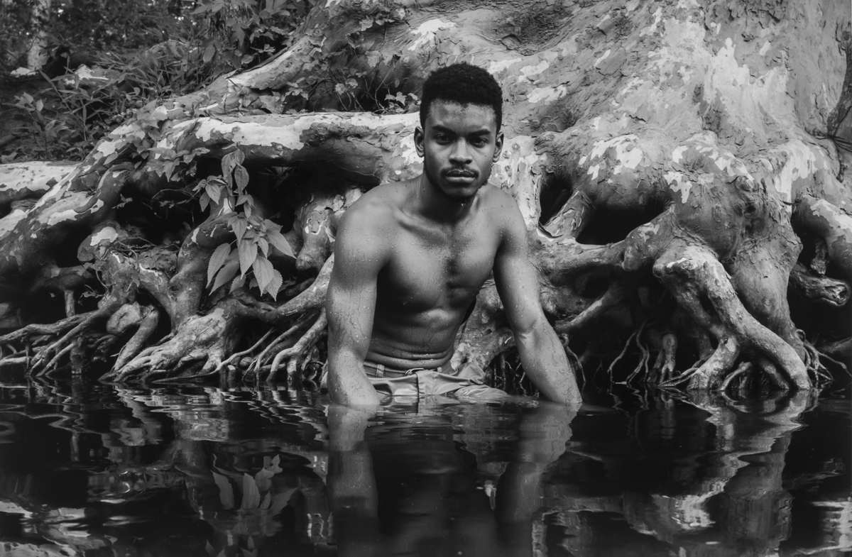 A grayscale photo of a person with a dark mustache and dark hair. The person is sitting in water next to some large tree roots and is not wearing a shirt.