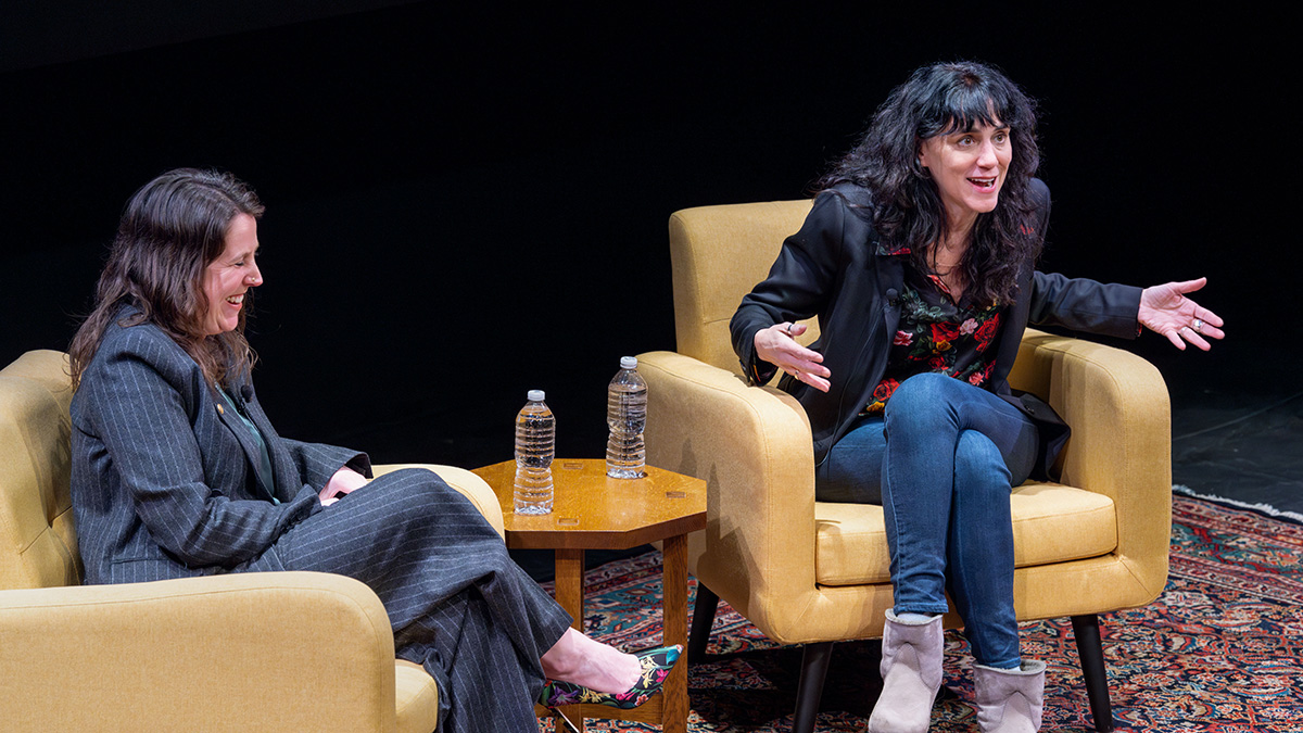 Two people sitting on a dark stage in large comfortable camel-tan chair speaking to each other and the audience.