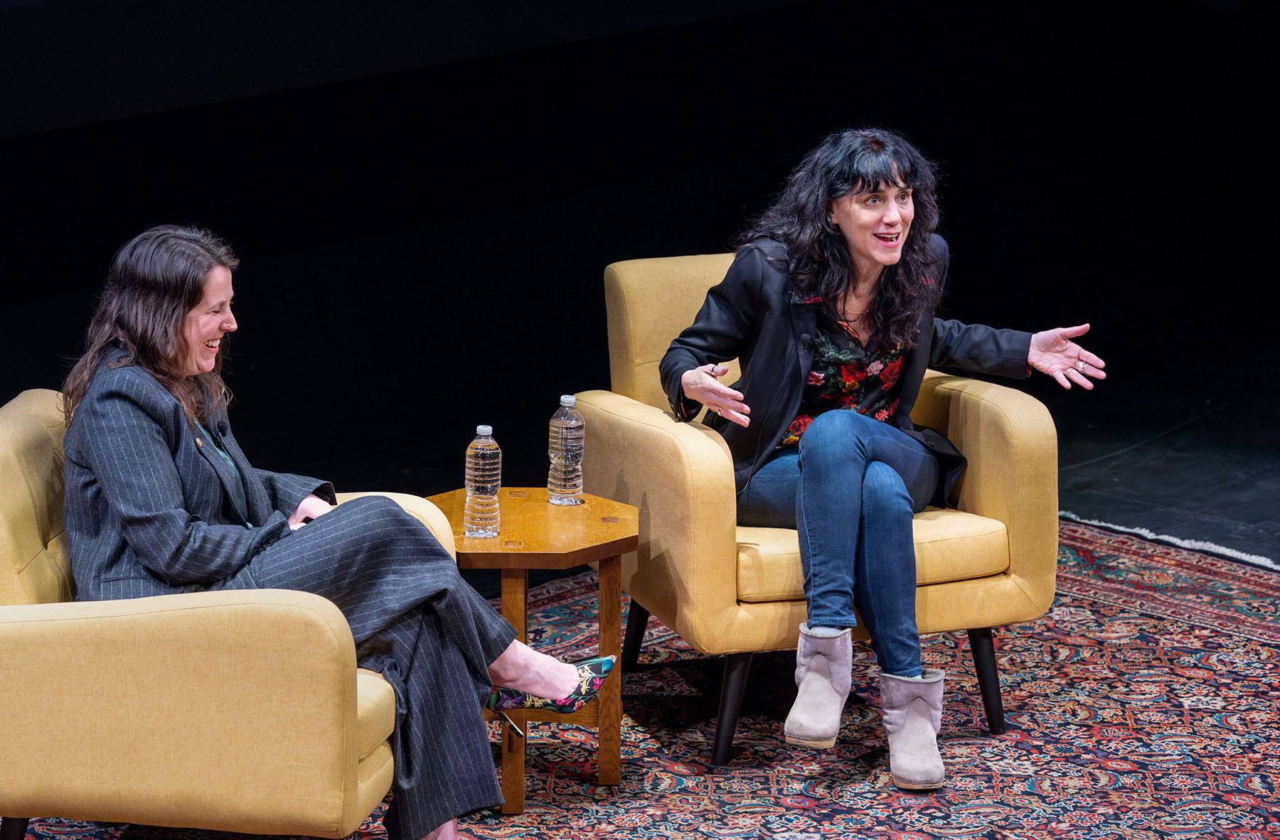 Two people sitting on a dark stage in large comfortable camel-tan chair speaking to each other and the audience.