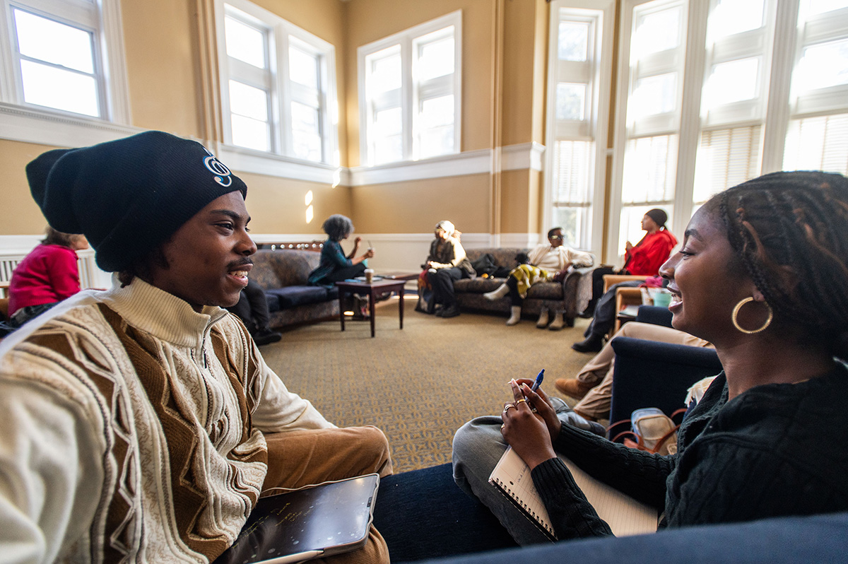 Two people sitting  and talking in a large room with bright windows with people seating in a circle around the room.