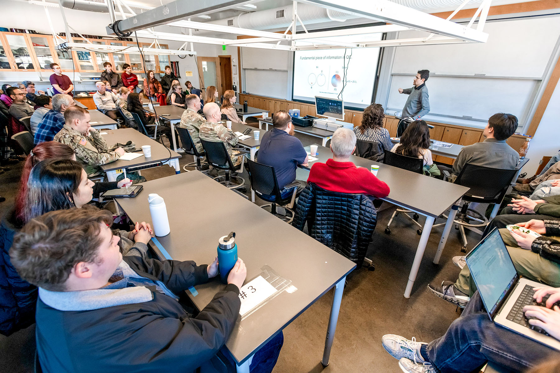 A large classroom with rows of tables with students sitting looking at the teacher and projector screen at the front of the room.