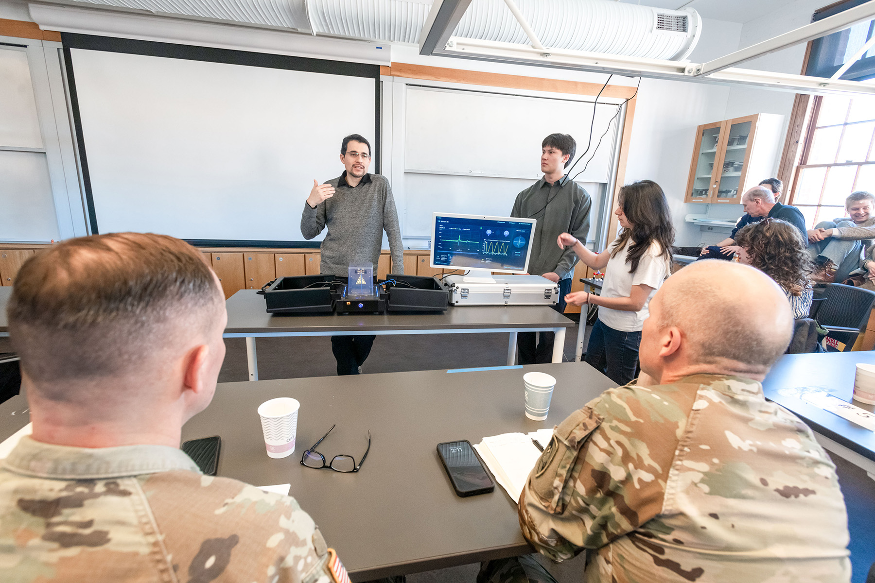 Military personnel in camouflage uniforms sitting in a classroom with a projector screen at the front with a teacher speaking.