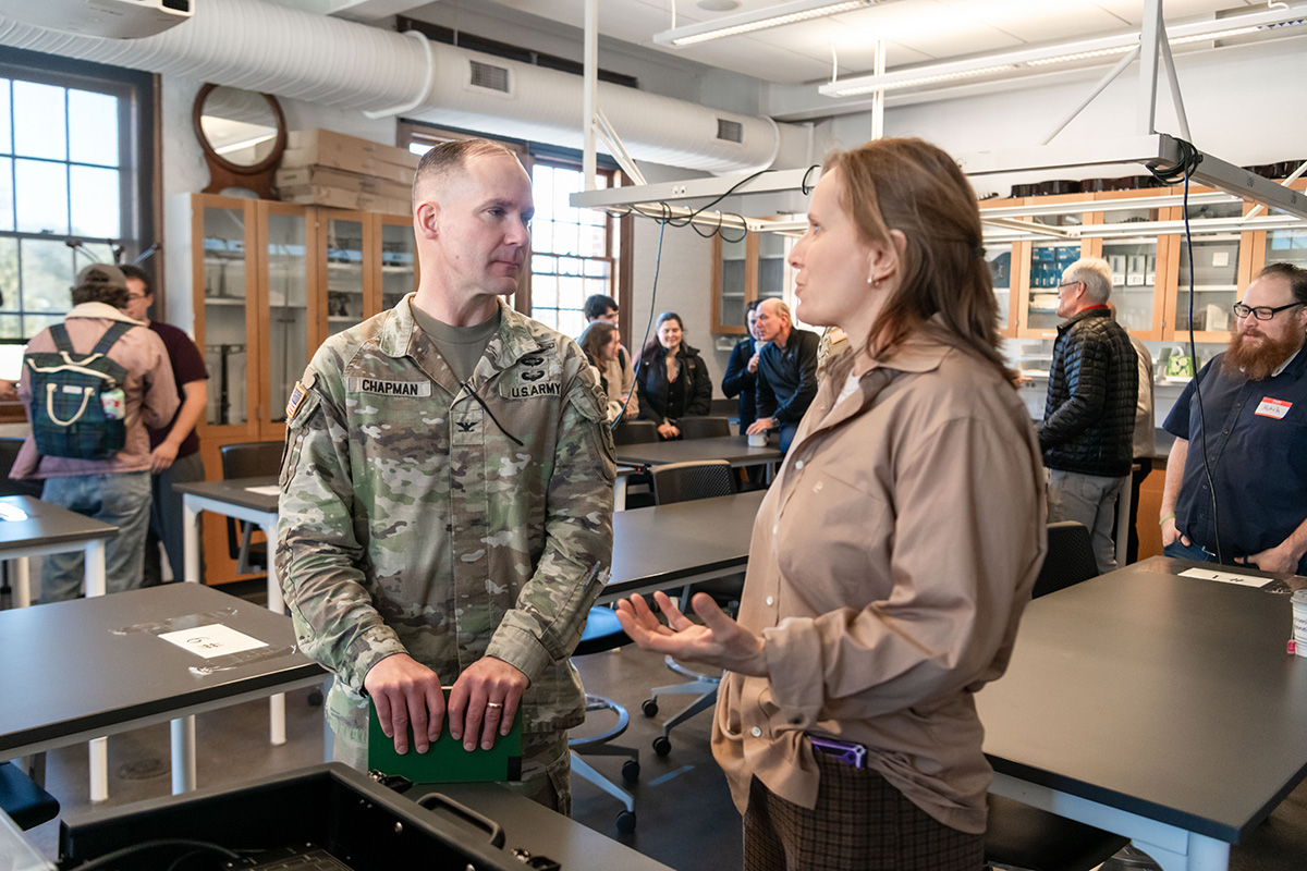 Two people standing in a classroom speaking to each other with other people and classroom tables in the background. One of the people speaking is wearing a camouflage, military uniform. 