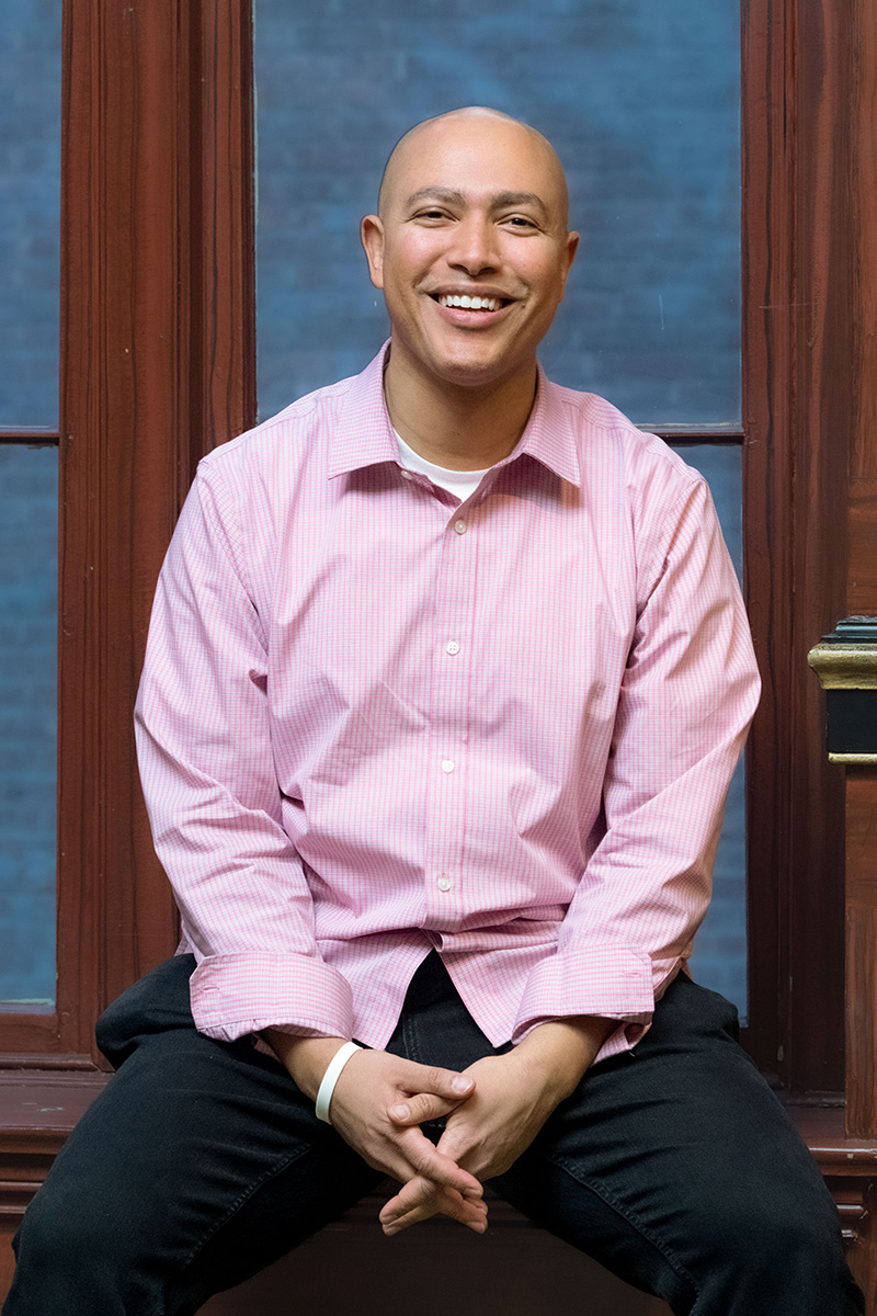 Person smiling and sitting on a stool in front of a dark-brown framed window wearing a button-down, light pink, collared shirt.