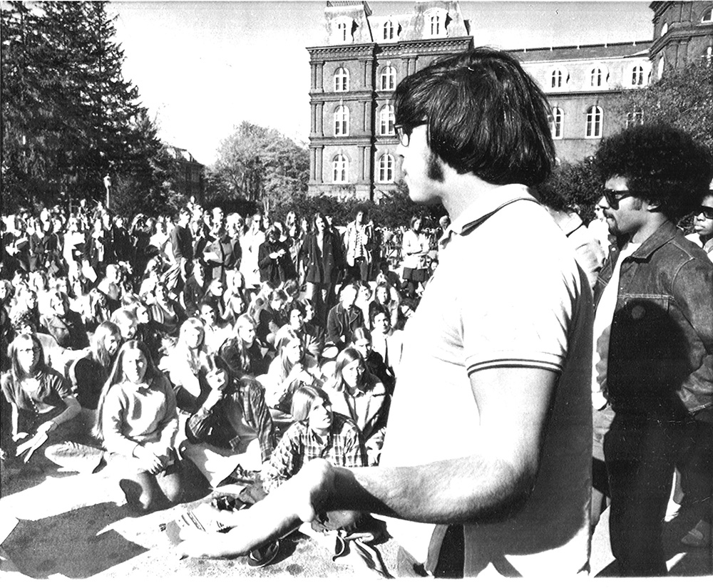 An old black and white image of a large gathering of people standing outside with Main building in the background.