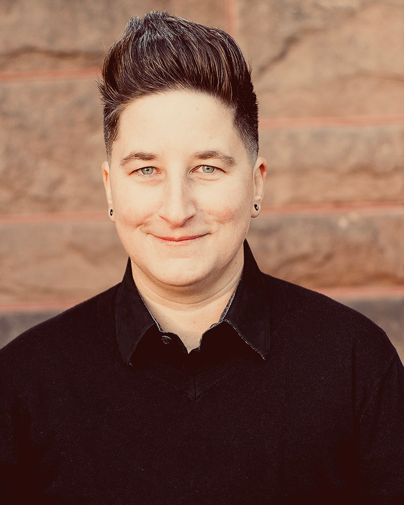 A portrait of a person in front of a red brick wall with tall brown hair and a black shirt.