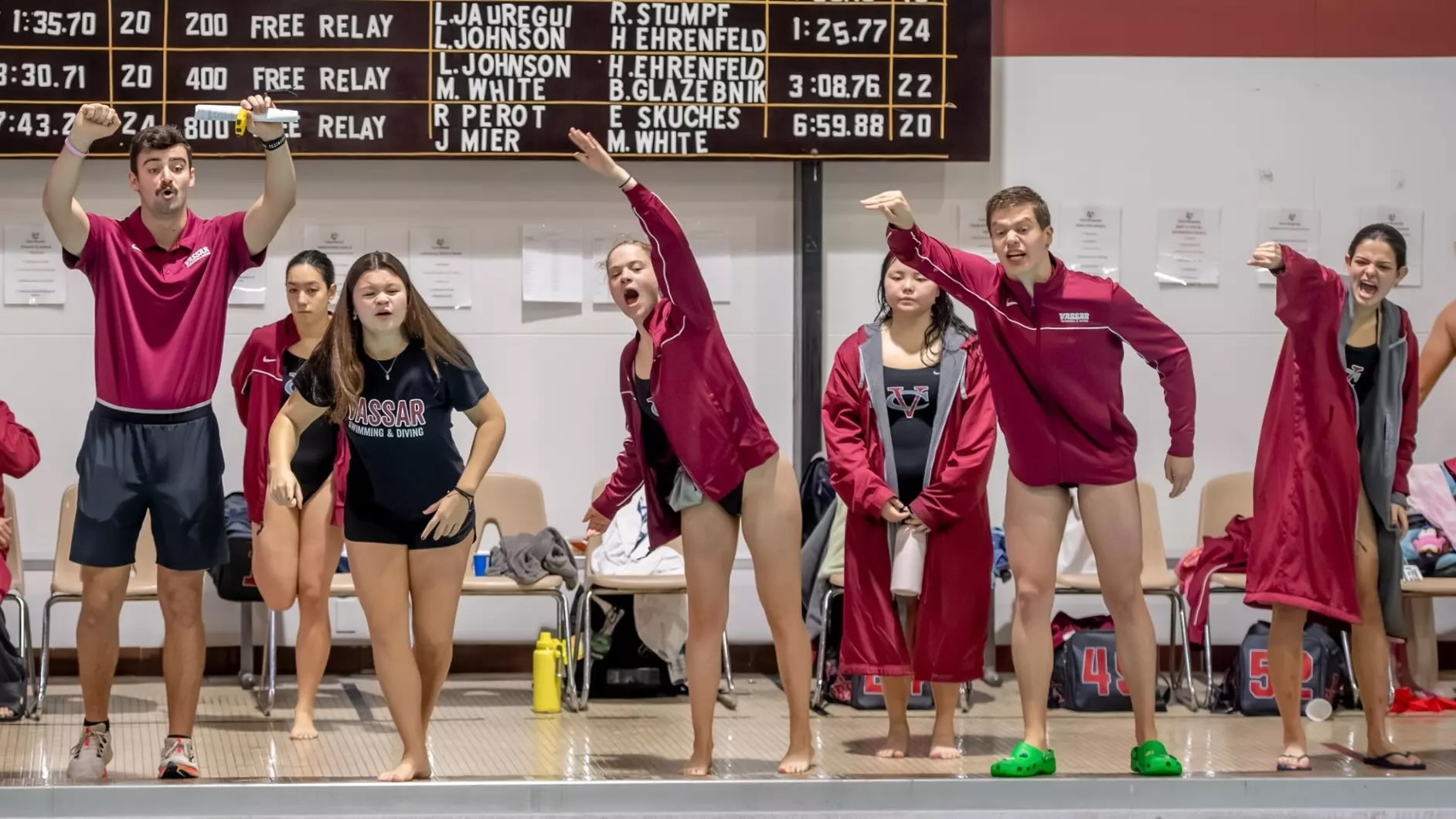 People in swimming gear standing poolside and cheering competitors off camera.