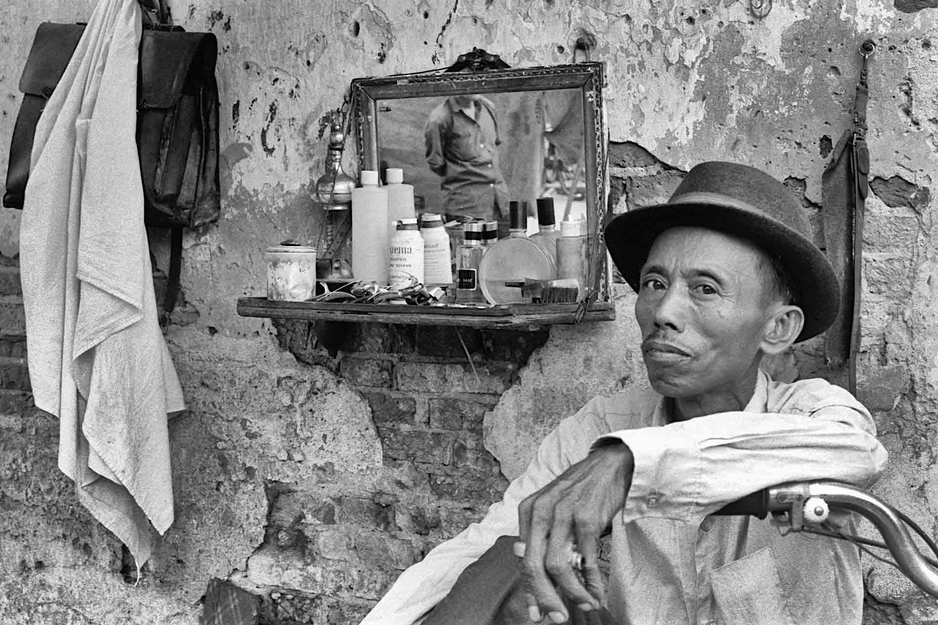 Seated barber set up on a sidewalk with a mirror hanging on a wall.