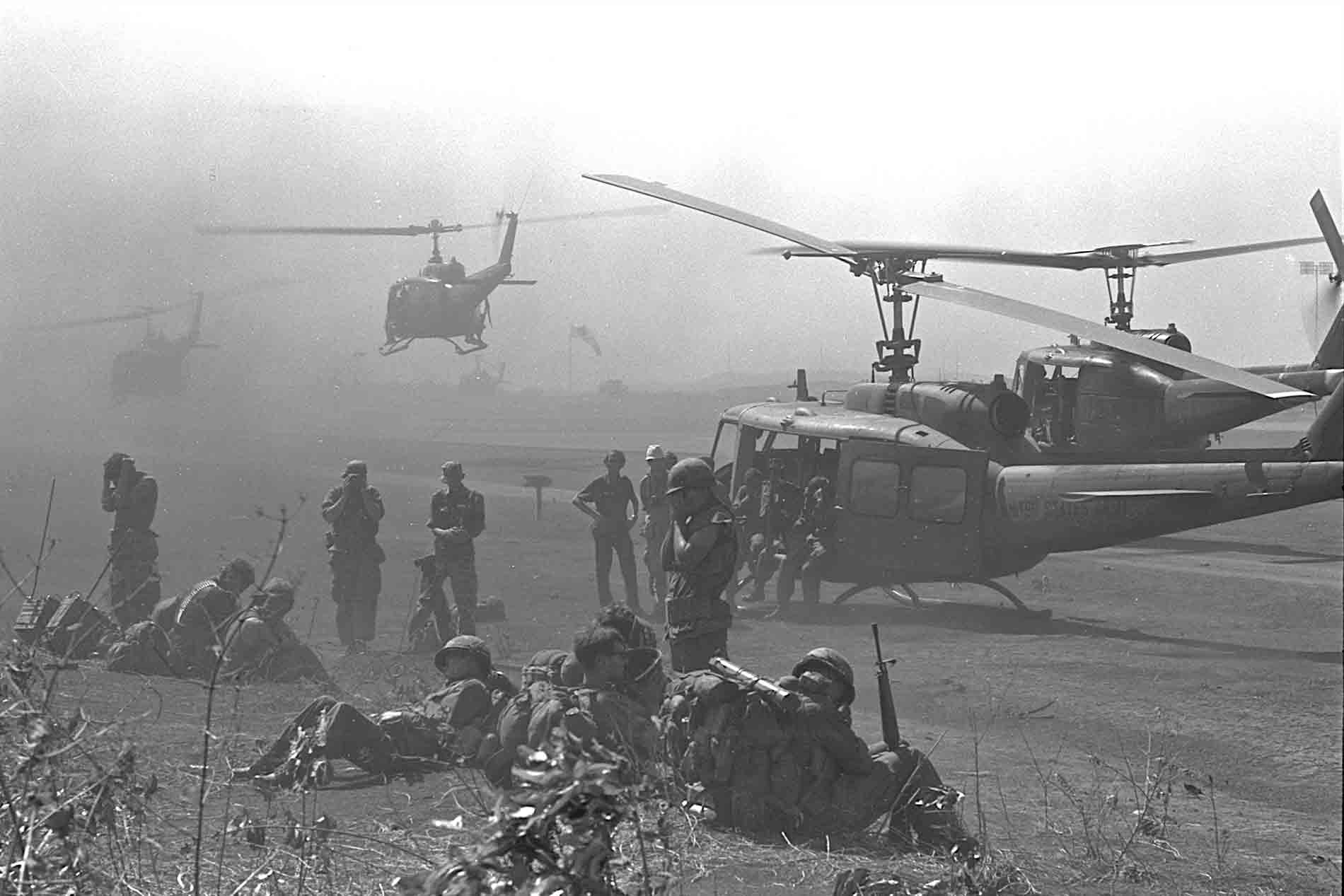 Helicopters landing in a field with troops standing and sitting nearby.