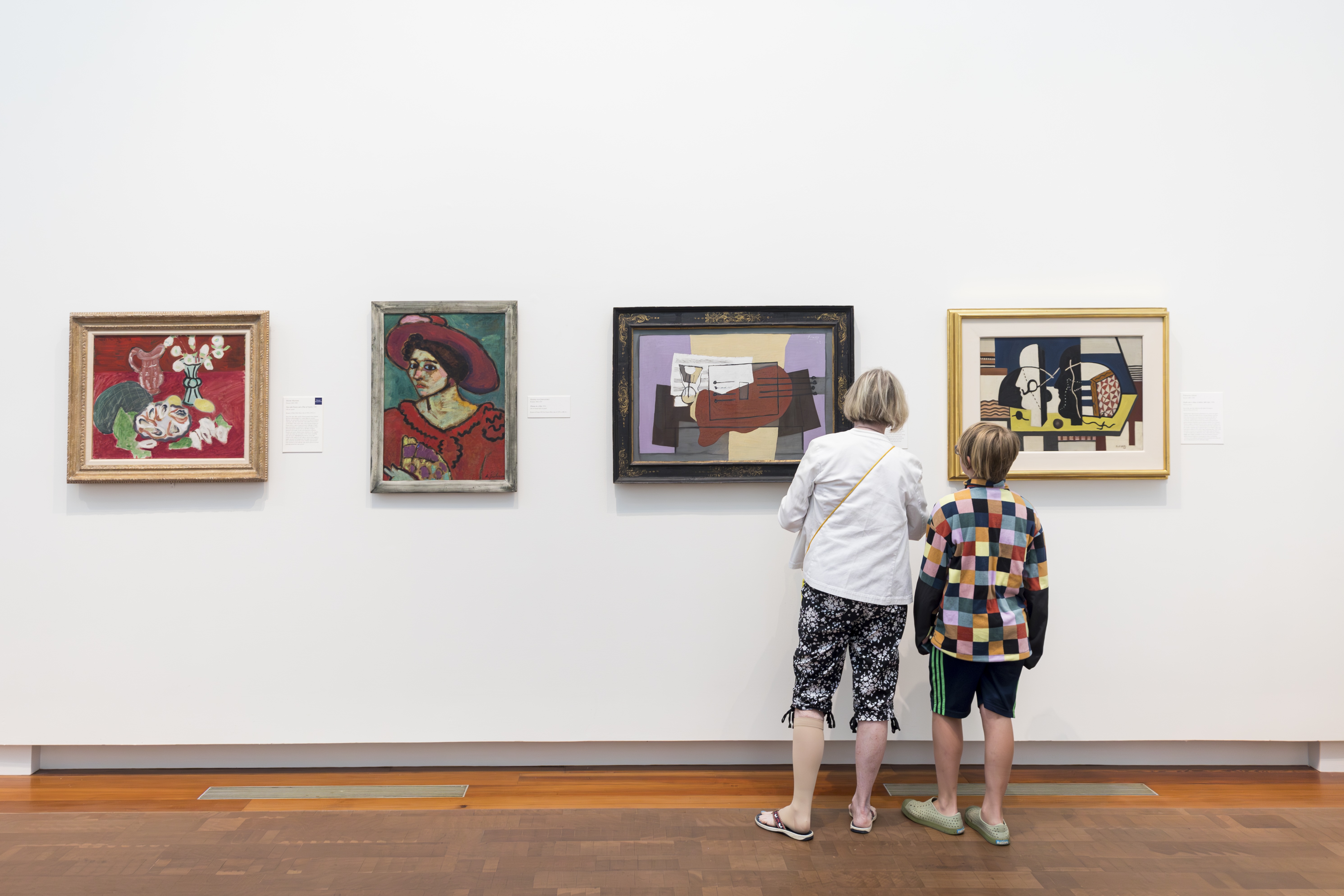 Adult woman and child stand with their backs to the viewer, looking at modern paintings in an art gallery.