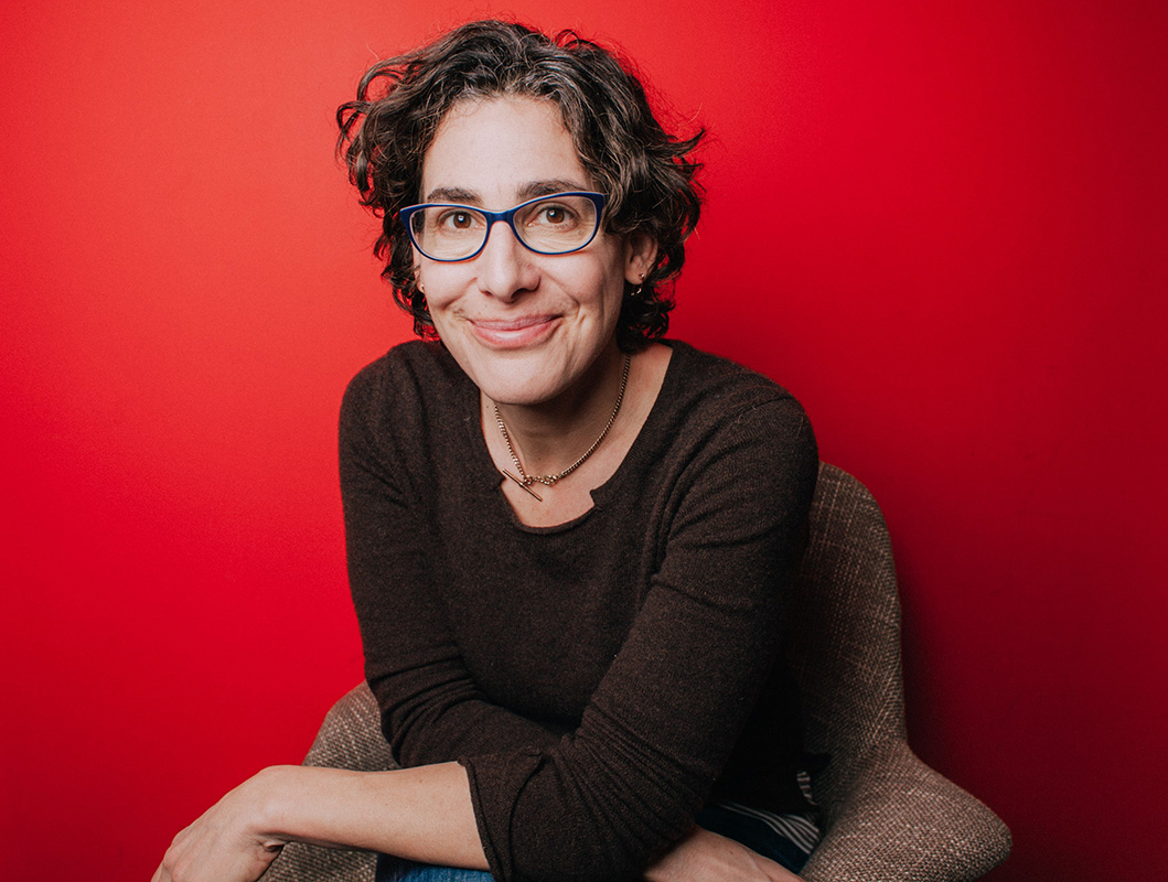 The subject is sitting in a light brown tweed chair, wearing a brown sweater, with a rich red background.