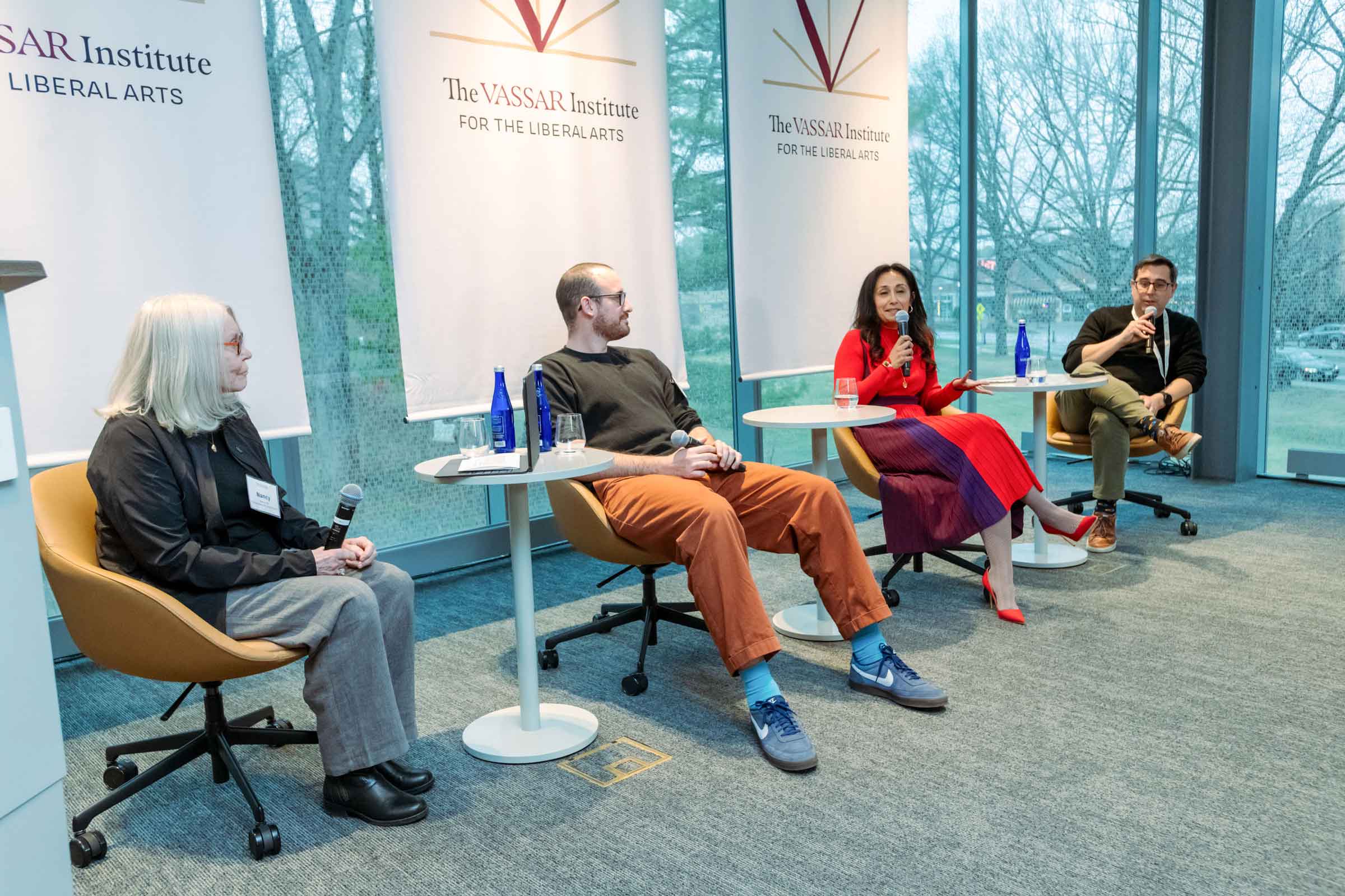 A seated panel of four people listen while one of the panelists holds and speaks into a microphone.