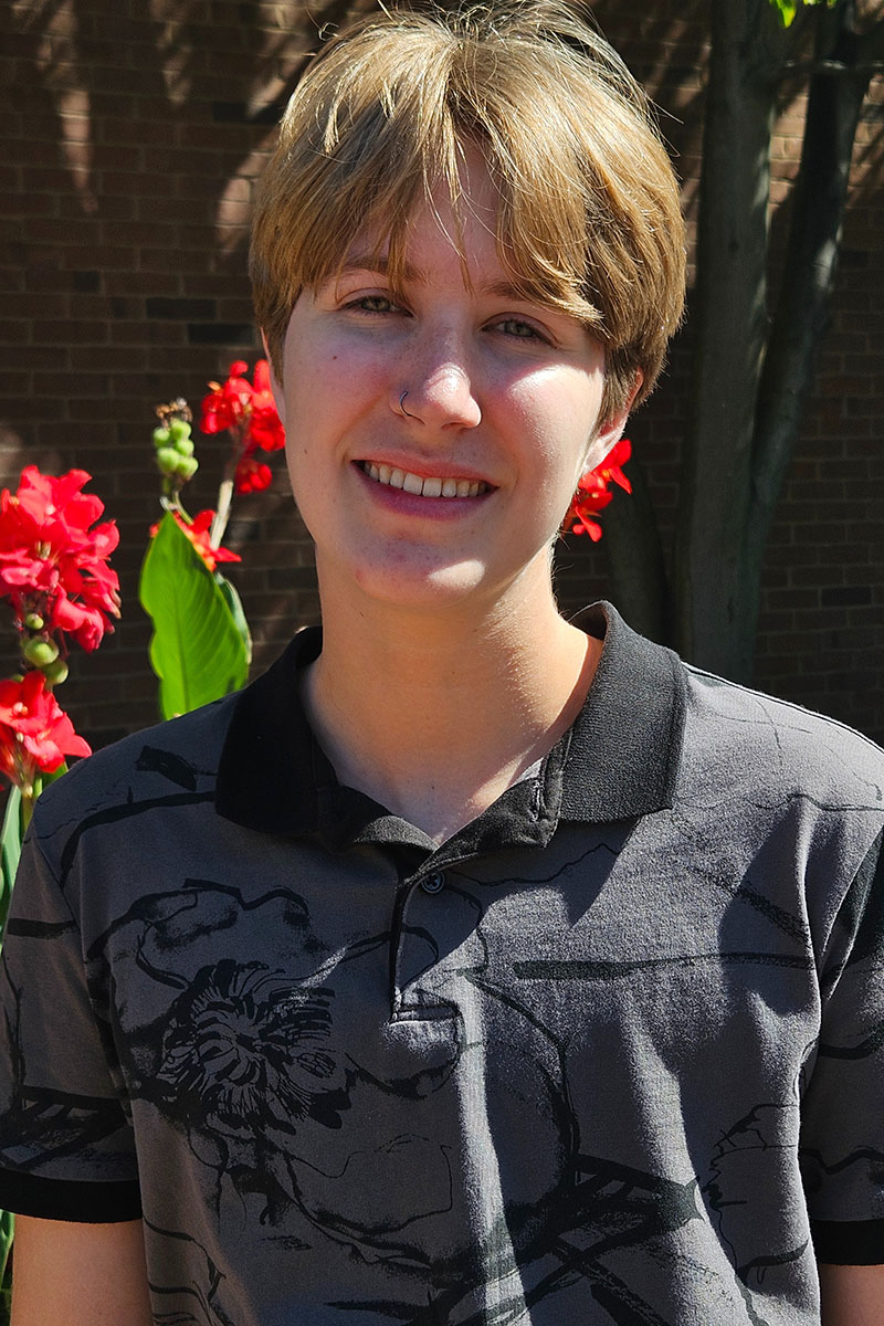 Person standing in sunlight with a relaxed smile and red flowers in background.