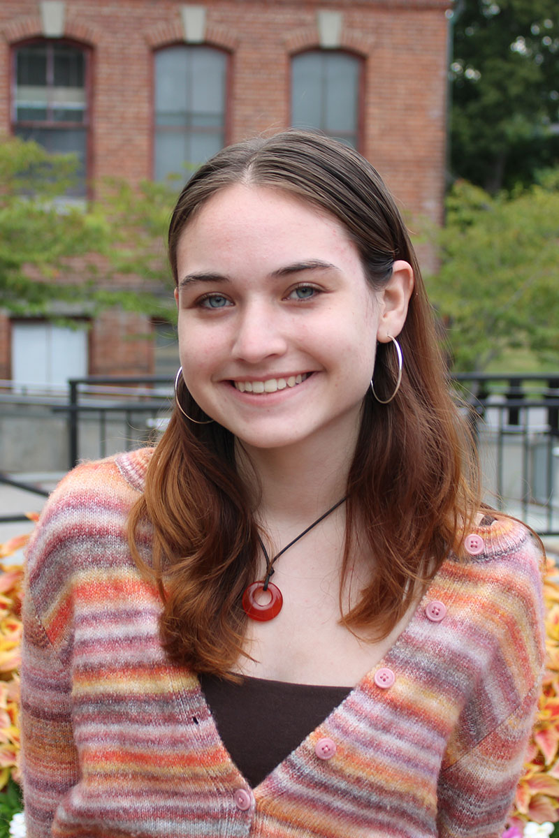 Person with brown hair and striped sweater smiles in front of flowers and brick wall.