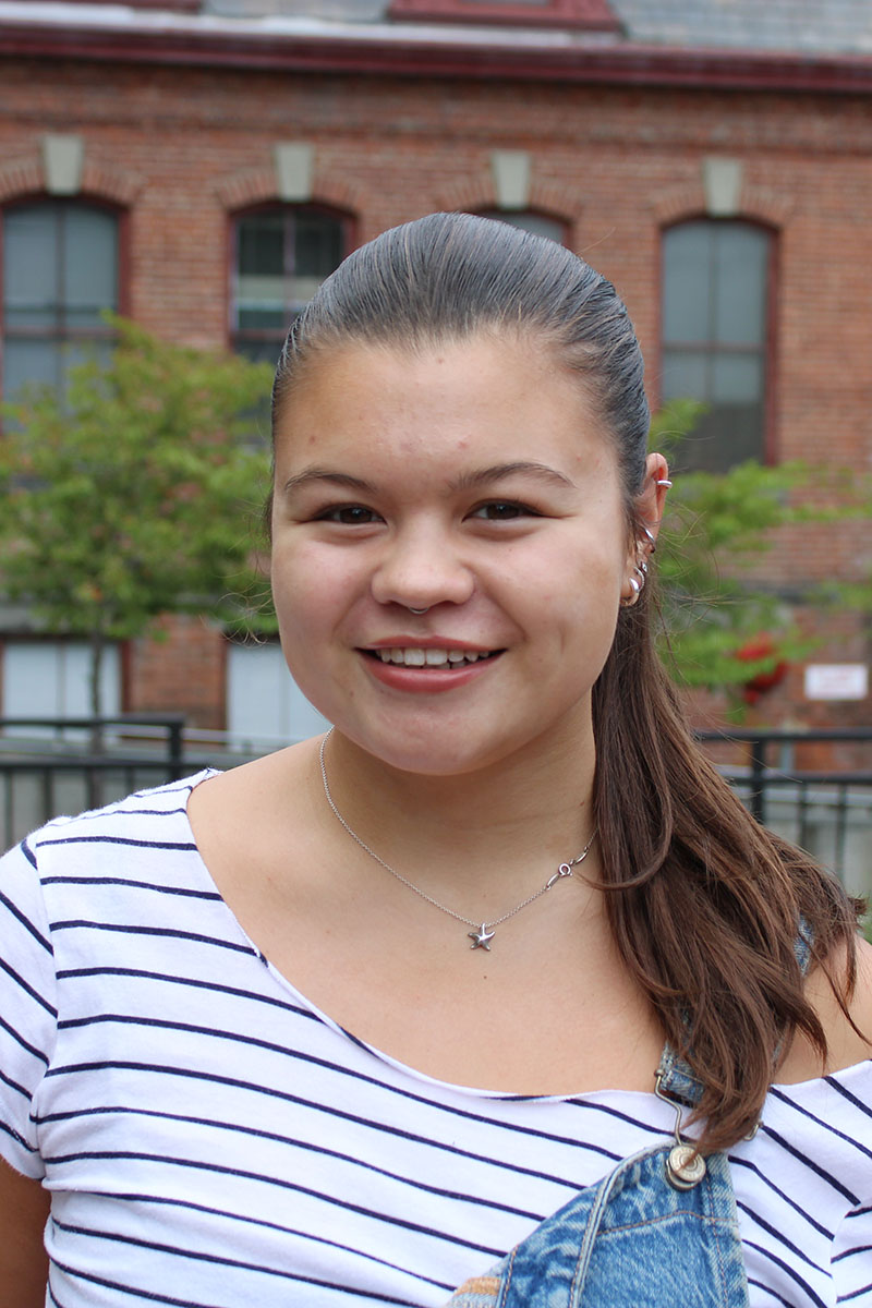 Person with hair pulled back smiles outdoors wearing a striped shirt and denim overalls.