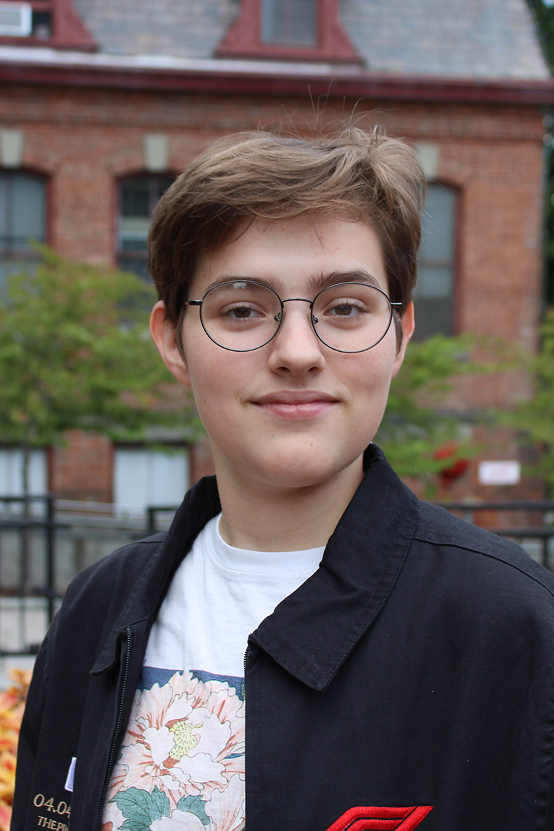 Person with short hair and glasses smiles outdoors in front of a brick building.