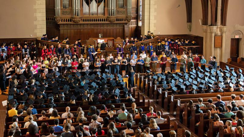 A large group of people sitting in a chapel. The people on stage are wearing different color graduation outfits while the people sitting at the pews are in traditional black graduation caps and gowns.