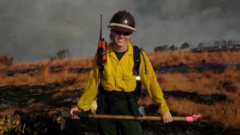 Pictured: Samantha Orient ’05. A person standing with fire-fighting gear (caution-yellow shirt and overalls) holding hatchet/shovel.