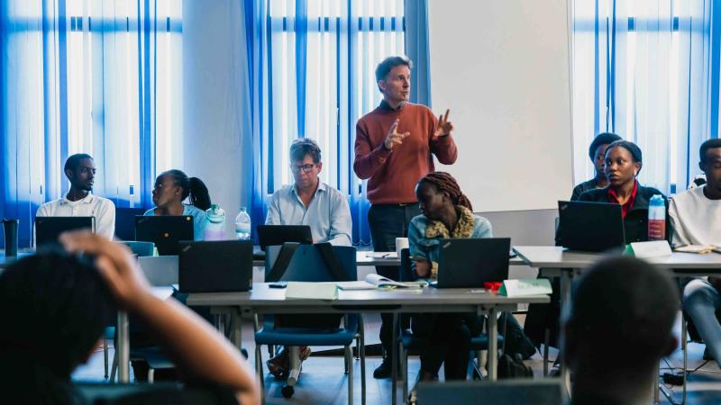 One person standing and speaking behind people listening and sitting at desks in a classroom setting with large windows and lots of natural light.